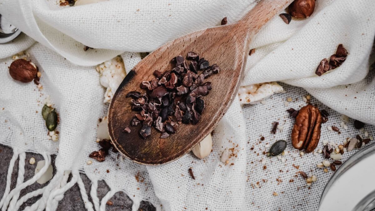 Top view of rustic wooden spoon with cacao nibs on white fabric surrounded by assorted nuts.