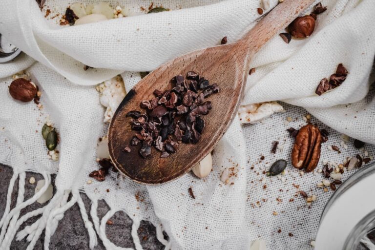Top view of rustic wooden spoon with cacao nibs on white fabric surrounded by assorted nuts.
